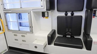 Crestline ambulance interior showing a rear-facing seat with integrated seatbelts, padded wall cushions, and an open storage drawer beneath the seat, alongside aluminum sliding cabinets and a yellow grab handle by the rear door