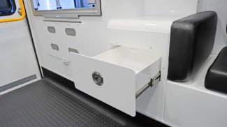 Interior view of a Crestline ambulance featuring an open drawer with a latch handle beneath a padded seat, next to lower wall storage compartments and a yellow grab handle by the rear door