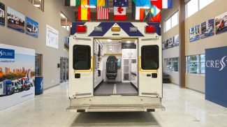 Rear view of a Crestline ambulance with both back doors open, showcasing a clean, organized interior equipped with seating, storage cabinets, and safety restraints, displayed inside the Crestline manufacturing facility
