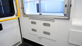 Interior side wall of a Crestline ambulance showing a lower storage access panel beneath aluminum sliding cabinets, with a yellow grab handle positioned next to the rear door