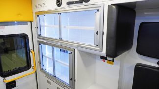 Interior view of a Crestline ambulance featuring upper and lower aluminum storage cabinets with sliding glass doors, black protective padding, and a yellow grab handle beside the rear entry door