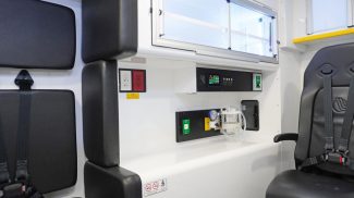 Patient compartment of a Crestline ambulance featuring a black action seat with harness, control panel with digital display, electrical outlets, oxygen controls, and onboard suction unit beneath overhead storage cabinets