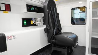 Crestline ambulance attendant seat equipped with an integrated 4-point safety harness, positioned beside the action wall with medical controls, oxygen outlets, and electrical panels