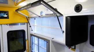 Illuminated upper storage cabinet inside a Crestline ambulance, featuring clear lift-up doors with gas struts, white shelving, and LED lighting above a secondary cabinet with sliding glass doors near the rear entrance