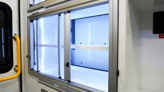 Interior view of an illuminated Crestline ambulance storage cabinet with clear sliding glass doors, white shelving, and integrated LED lighting next to a rear window and yellow grab handle