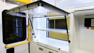 Open interior storage cabinet inside a Crestline ambulance module, featuring bright LED lighting, white shelving, and a clear lift-up door supported by gas struts next to a rear window with a yellow safety grab handle