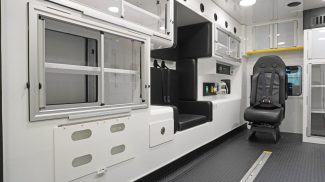 Interior of a Crestline ambulance featuring clean white cabinetry with sliding glass doors, padded seating with safety restraints, integrated control panels, and a durable non-slip floor designed for efficient patient care and crew safety