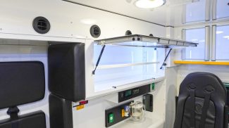 Crestline ambulance interior featuring an open upper storage cabinet with LED lighting, padded seating with restraints, air vents along the wall, and a mounted oxygen system below the work counter