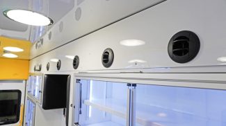 Close-up of the Crestline ambulance ceiling and upper wall showing round air vents, bright LED lighting, and illuminated storage cabinets with sliding glass doors