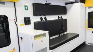 Crestline ambulance interior showing a black bench seat with seatbelts, overhead storage cabinets, a control panel, and a waste bin compartment built into the side cabinet