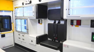 Crestline ambulance interior featuring a black padded attendant seat with seatbelts, overhead and side storage compartments with sliding doors, and electrical outlets integrated into the wall panel