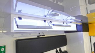 Overhead storage cabinets inside a Crestline ambulance with clear lift-up doors, interior lighting, and a black padded bench seat below