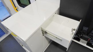 Interior view of a Crestline ambulance showing a white cabinetry unit with an open drawer beside a black seat equipped with safety restraints