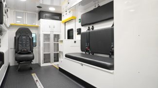 Interior view of a Crestline ambulance showing a clean, organized patient care area with black seating, 4-point seatbelts, upper and lower storage cabinets, and a durable non-slip diamond plate floor