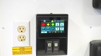 Close-up of a Crestline ambulance interior control panel displaying lighting and temperature settings beside a dual 120V electrical outlet with a yellow safety notice label