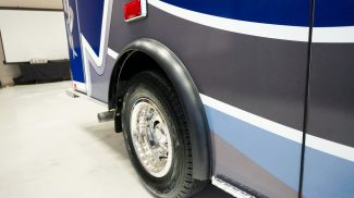 Close-up of a Crestline ambulance rear wheel with chrome hubcap and blue and gray body graphics, parked indoors on a smooth concrete floor