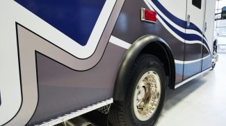 Close-up of a Crestline ambulance side panel showing blue and gray graphics, rear wheel with chrome hubcap, and visible exhaust pipe below the diamond plate trim