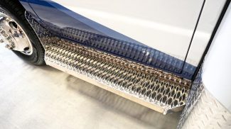 Close-up view of a Crestline ambulance side step with diamond plate aluminum and textured grip surface, showing part of the wheel and lower body panel with blue graphics