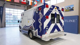 Rear view of a Crestline CCL 166 ambulance inside a showroom, showcasing its bold blue and white chevron design, rear entry doors, and diamond plate step bumper