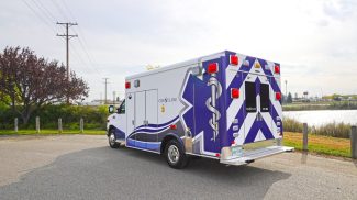 Rear and side view of a Crestline CCL 166 ambulance parked near a waterfront, showcasing its bold blue and white chevron design and Star of Life graphics