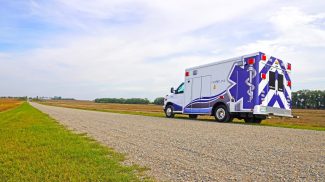 Crestline ambulance parked on a rural gravel road beside open fields under a bright, partly cloudy sky