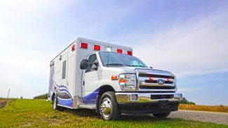 Low-angle view of a Crestline ambulance with emergency lights activated, parked on a grassy roadside under a bright blue sky