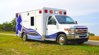 Side and front view of a Crestline ambulance with emergency lights activated, parked on a grassy roadside under a bright sky