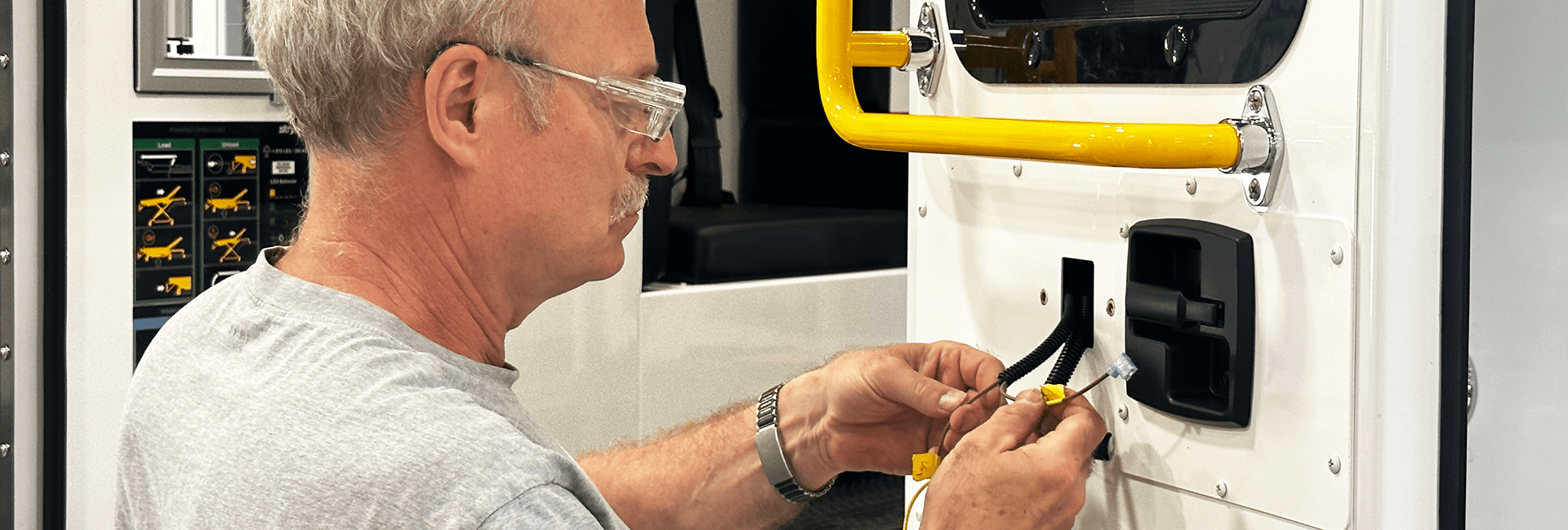 Technician wearing safety glasses works on wiring inside a Crestline ambulance, connecting components near the rear door with a yellow grab handle visible.