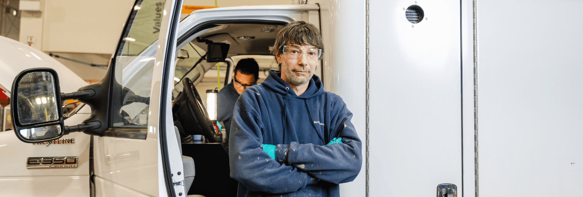 Factory technician standing beside a Crestline E-Series ambulance during assembly, with another team member working inside the cab in the background.