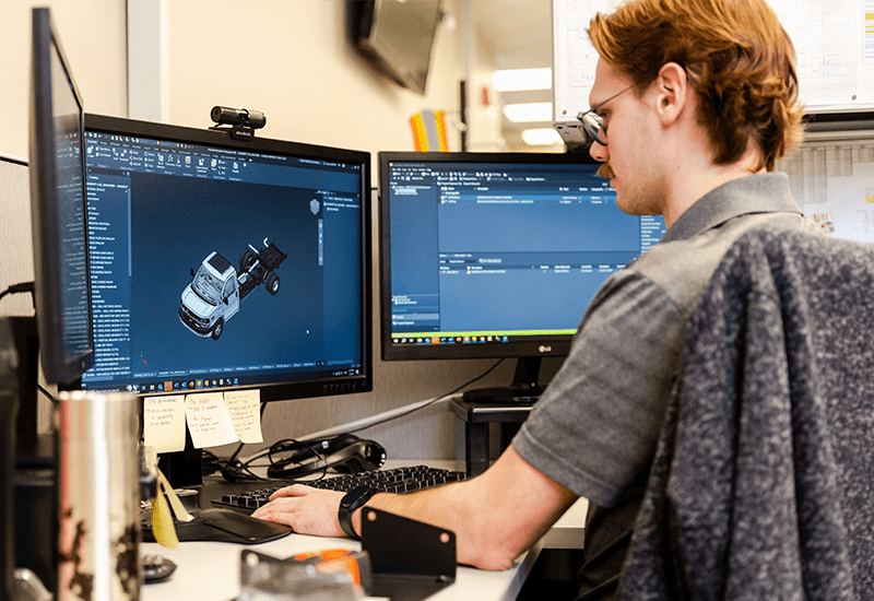 Crestline engineer works at a dual-monitor workstation designing a vehicle model in 3D software, focusing on the layout of an ambulance chassis
