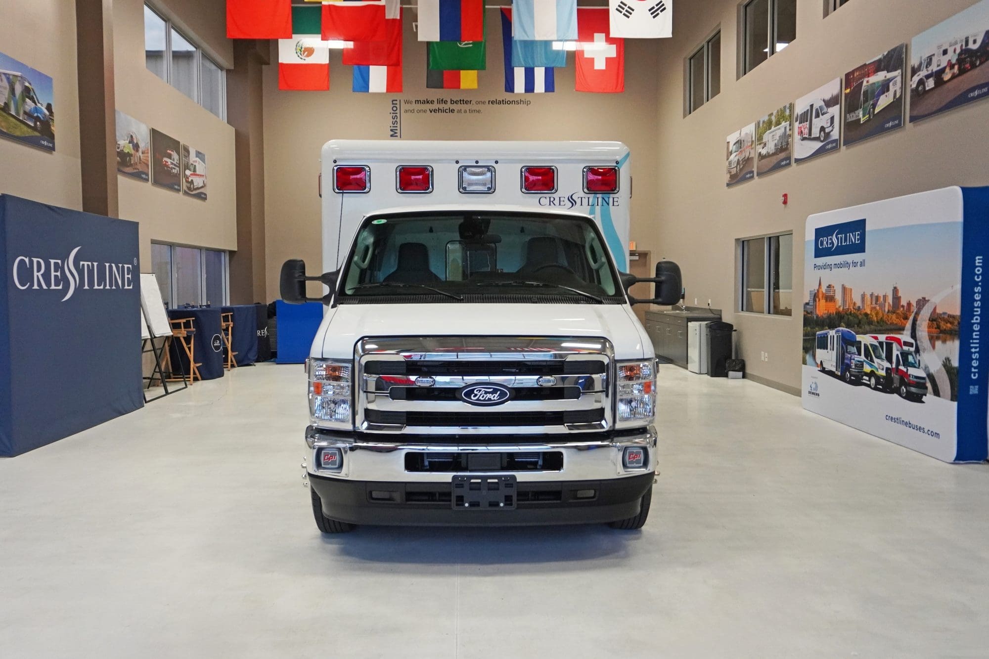Front view of a Crestline CCL 166 ambulance built on a Ford chassis, displayed inside the Crestline manufacturing facility with branding and international flags in the background
