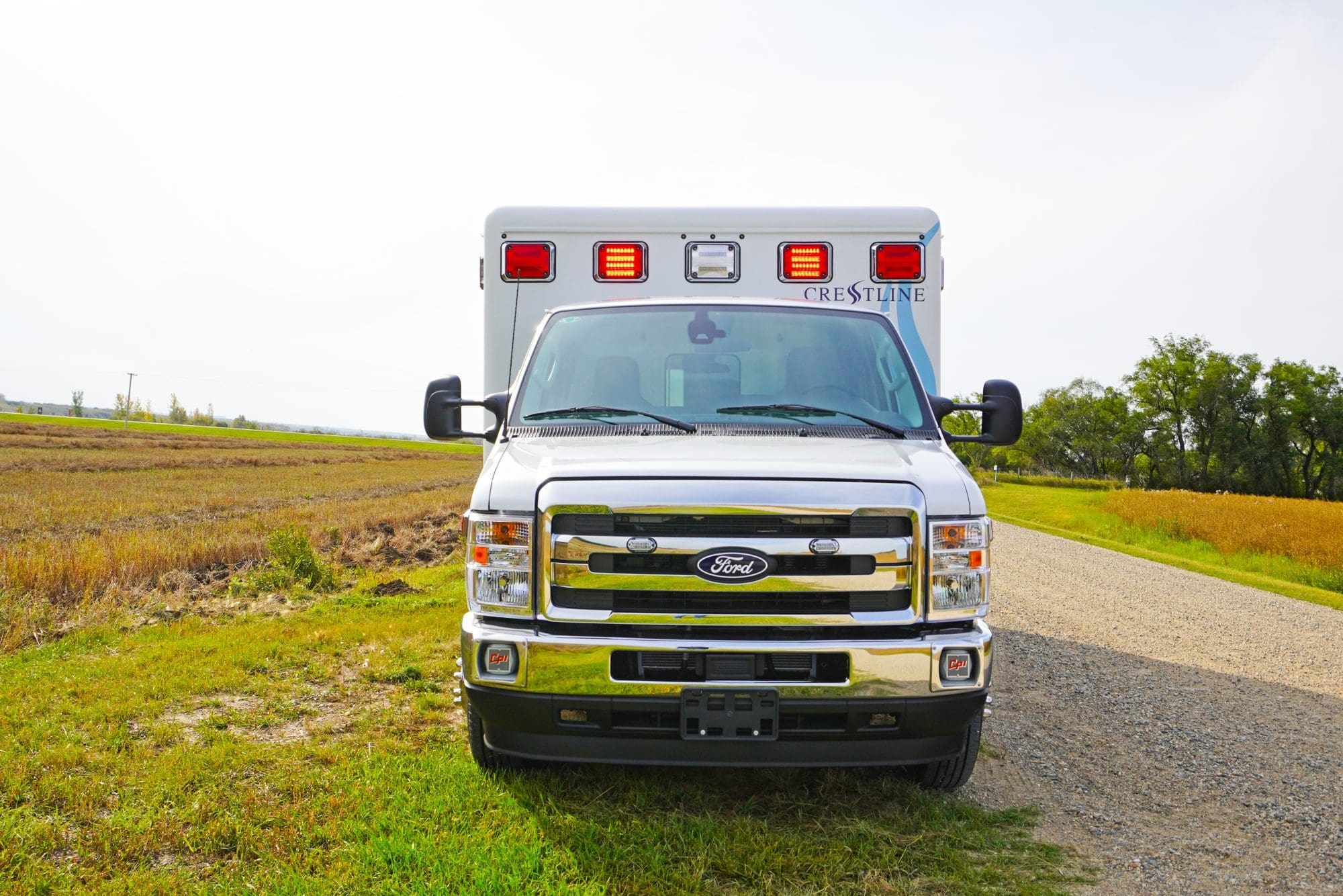 Front view of a Crestline ambulance with red emergency lights on, parked on a gravel road beside an open rural field