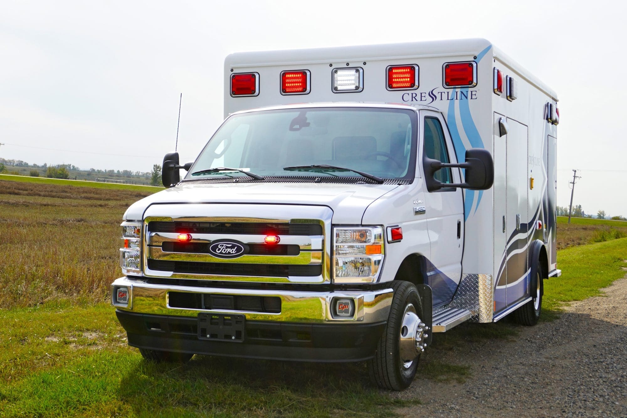 Front view of a Crestline ambulance with emergency lights illuminated, parked on a rural roadside surrounded by open fields