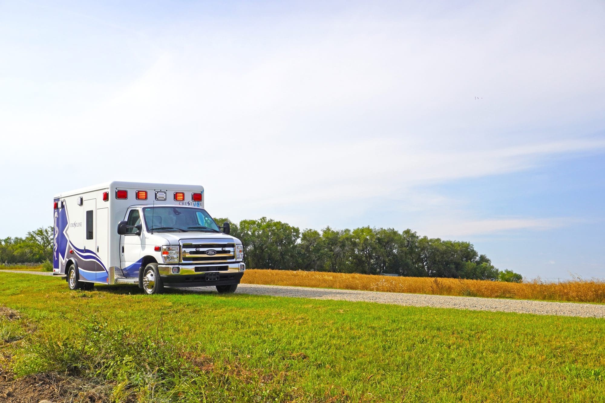 Crestline ambulance on the side of a rural gravel road surrounded by green grass and open fields under a bright blue sky