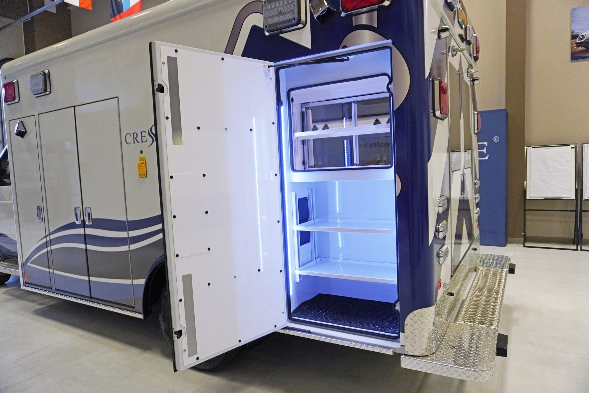 Open rear-side compartment of a Crestline ambulance illuminated with bright LED lighting, showcasing organized interior shelving and durable diamond-plate step surfaces