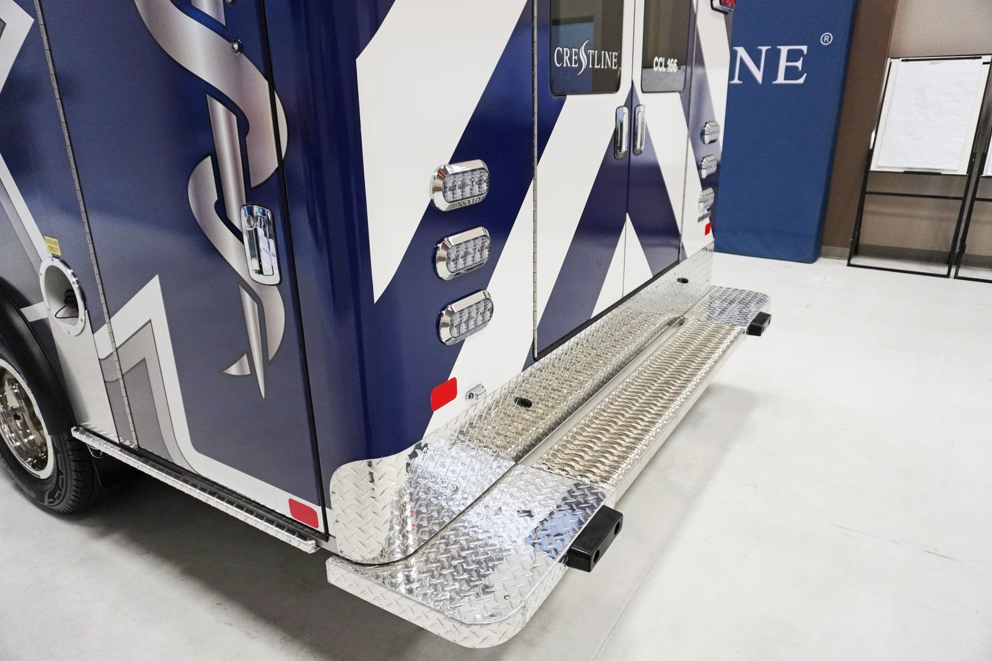 Close-up of the rear exterior of a Crestline CCL 150 ambulance, showing the diamond-plate rear step, bright LED lighting, and sleek blue-and-white graphics featuring the Star of Life design