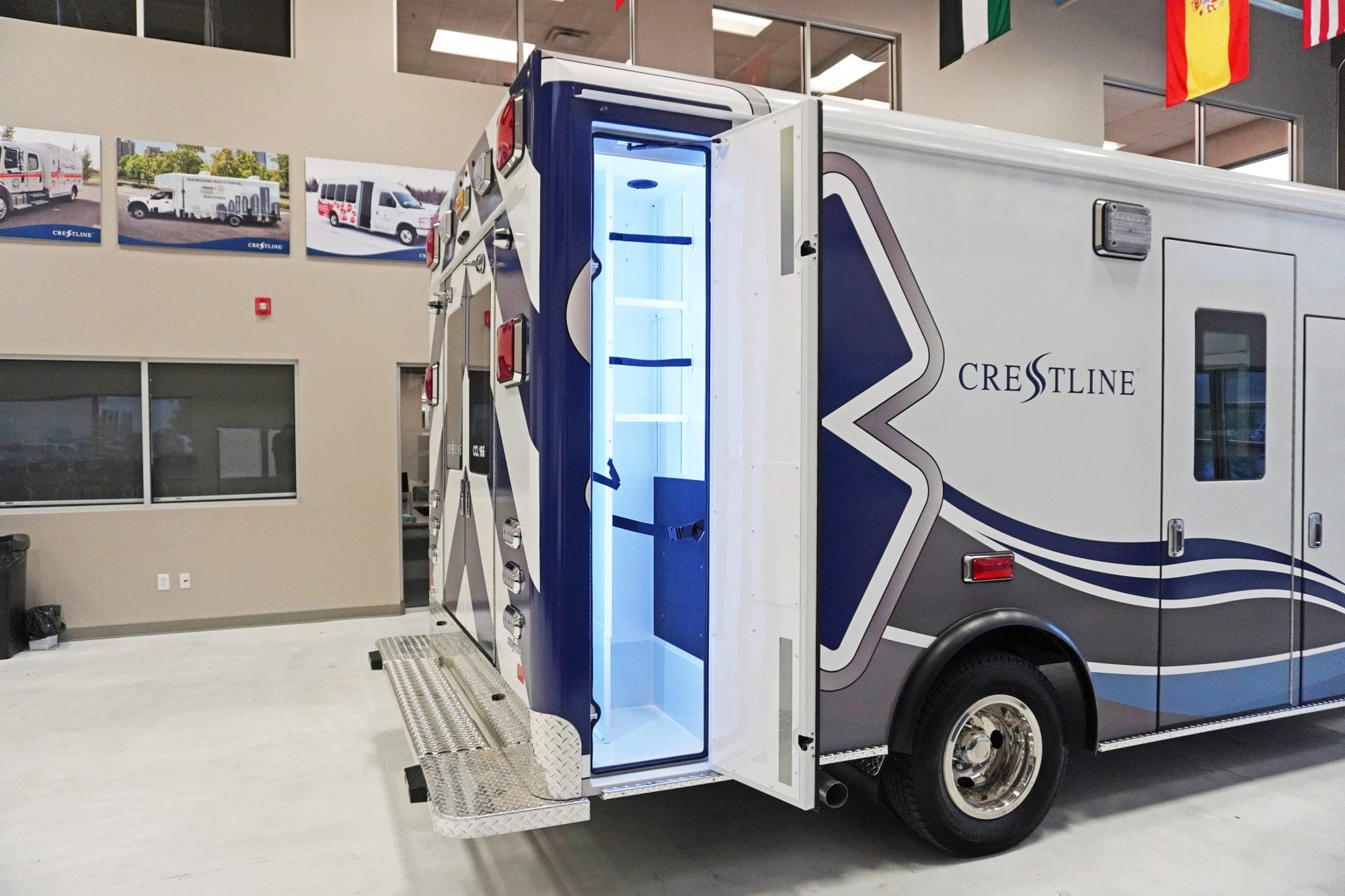 Rear-side view of a Crestline ambulance with an open exterior compartment illuminated in blue light, showing interior shelving and storage space