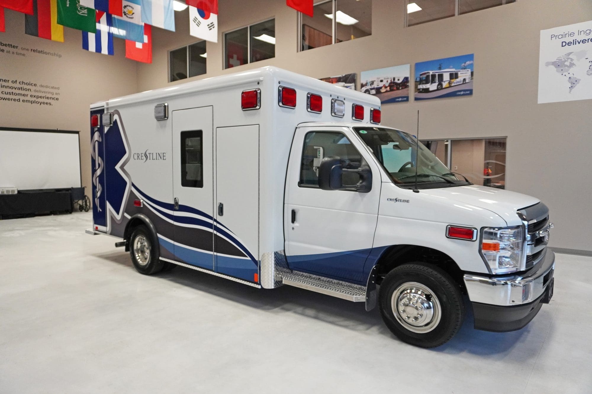 Crestline CCL 166 ambulance shown in a showroom, featuring blue and gray wave graphics along the sides and a large Star of Life design on the rear module