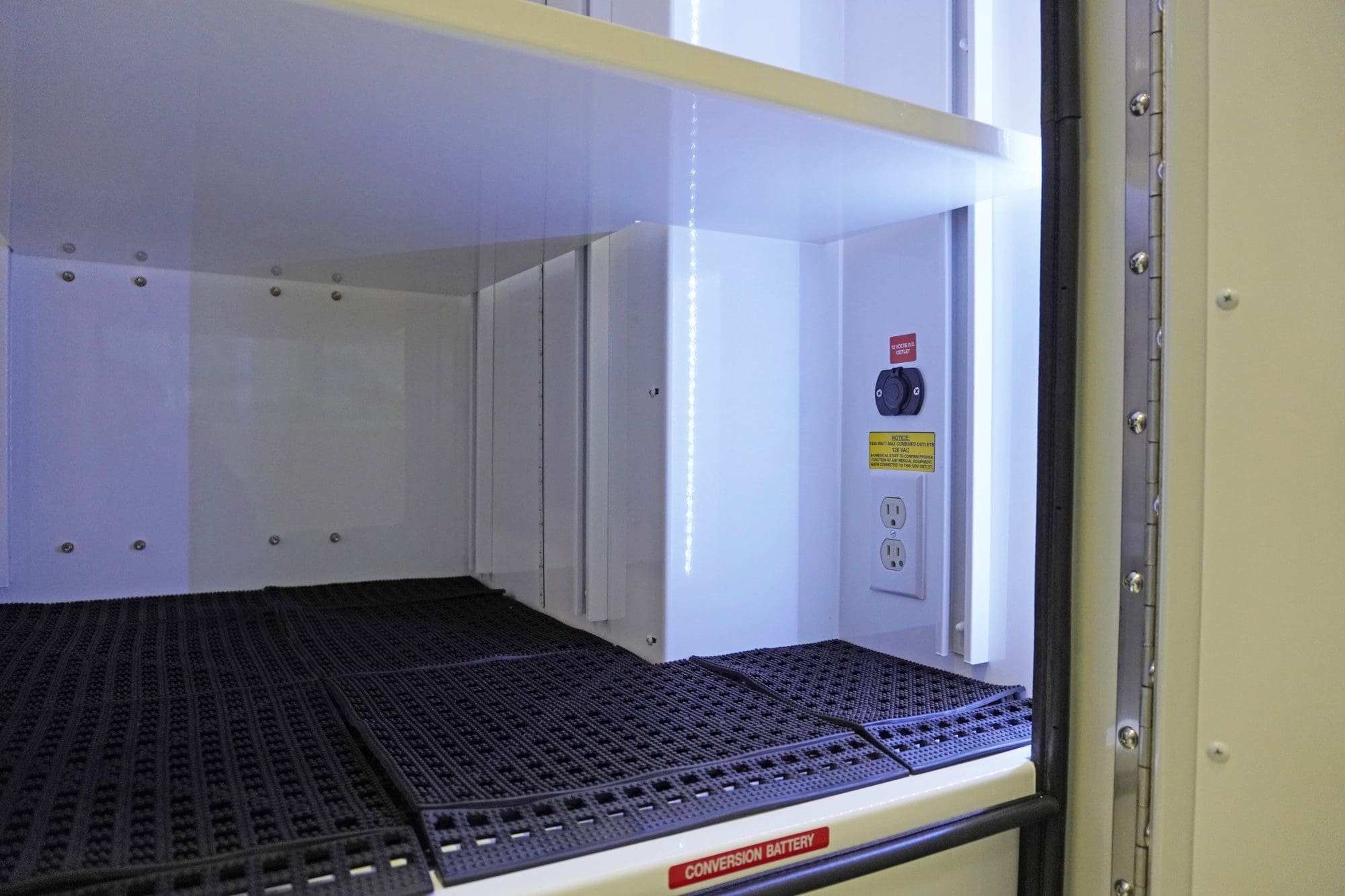 Close-up of an illuminated interior Crestline ambulance cabinet with textured black shelf mats, electrical outlets, and a labeled conversion battery switch
