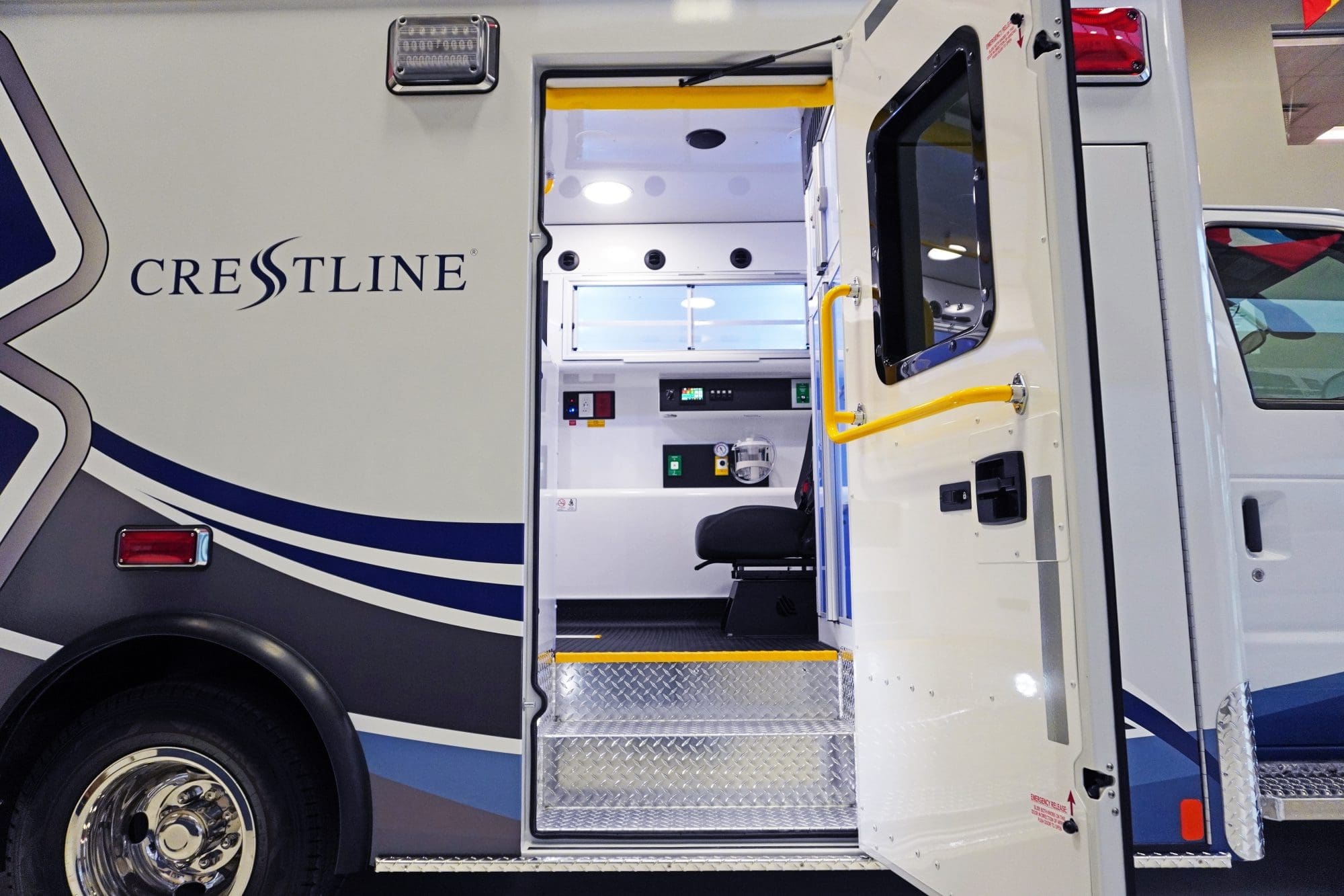 Open side door of a Crestline ambulance revealing the illuminated interior with black seating, control panels, and diamond plate entry steps