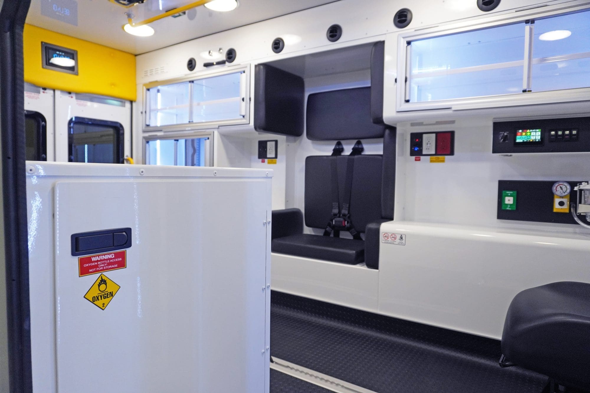 Interior of a Crestline ambulance featuring black seating with seatbelts, upper and lower storage cabinets, oxygen access panel, and control switches