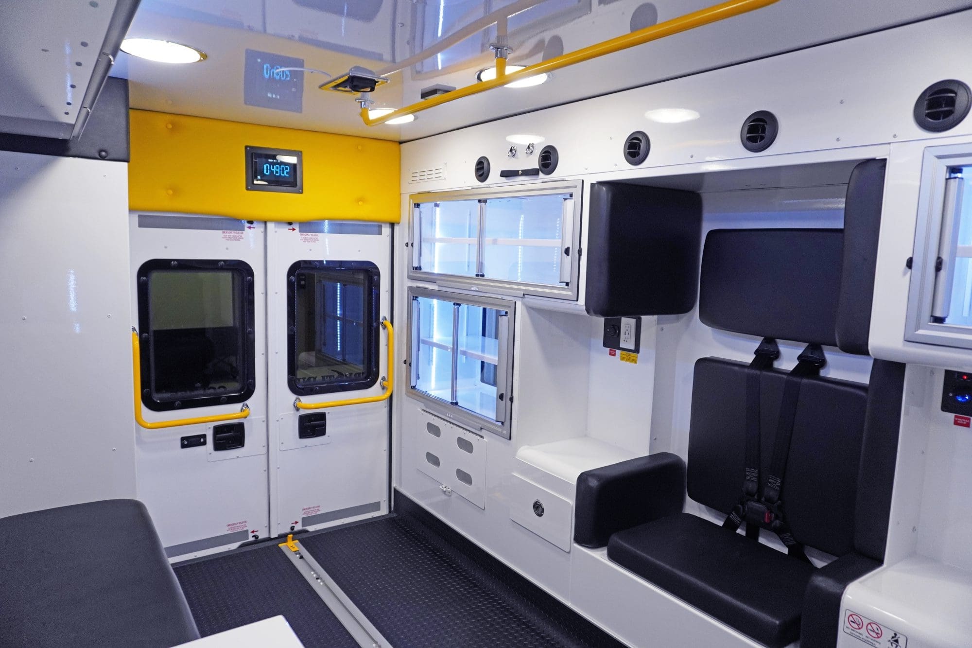 Crestline interior view of an ambulance showing rear entry doors with yellow grab handles, black seating with seatbelts, overhead lighting, and illuminated storage cabinets