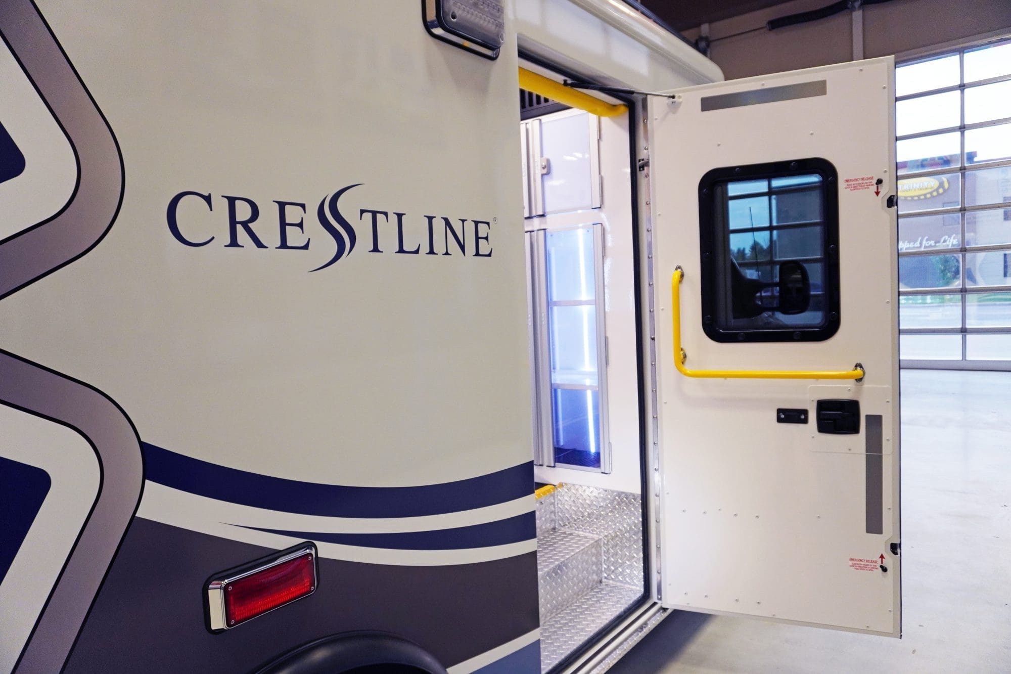 Side view of a Crestline ambulance with the entry door open, revealing bright yellow grab handles, diamond plate steps, and illuminated interior cabinets