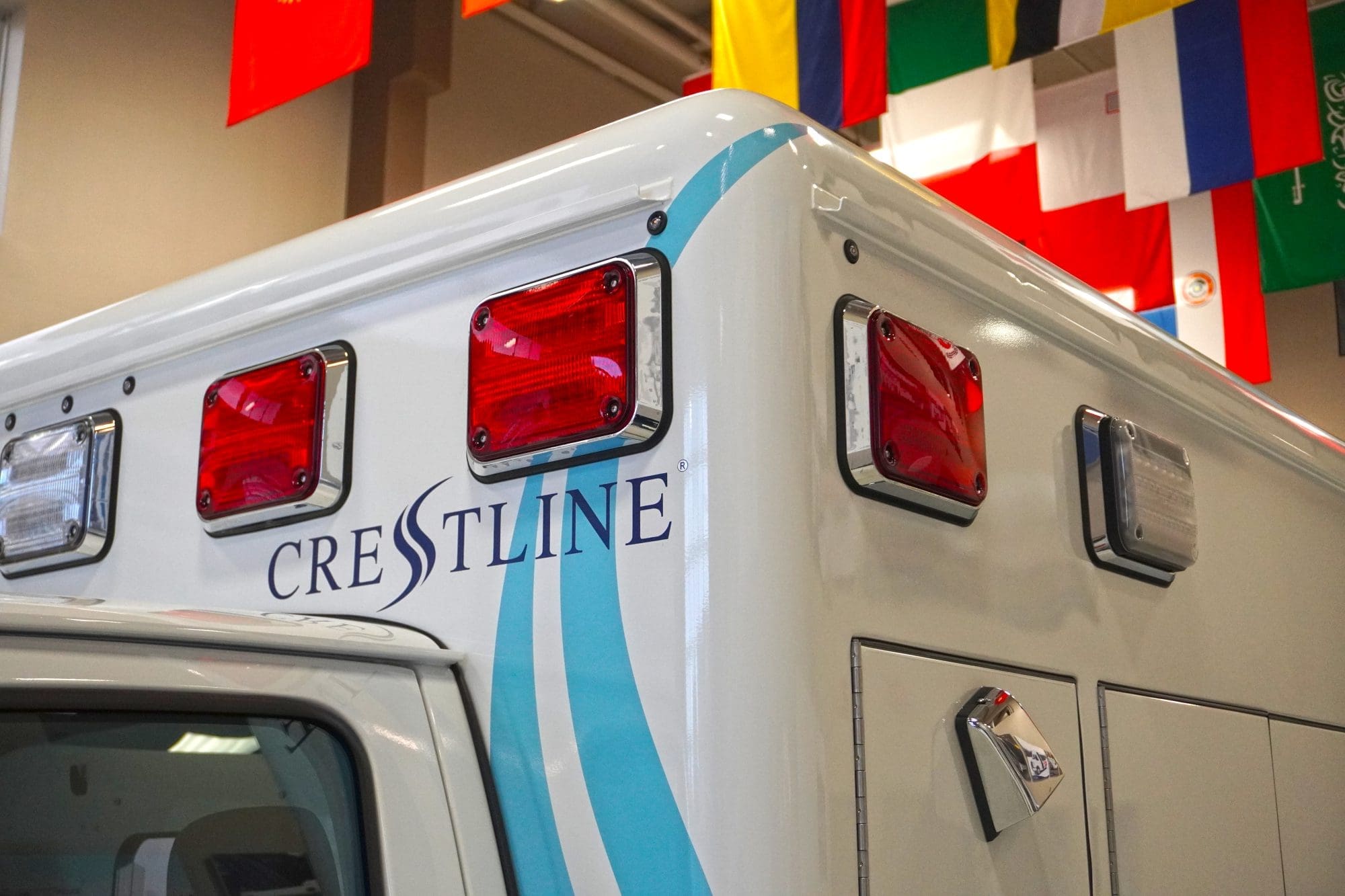 Close-up of the upper rear corner of a Crestline ambulance featuring red and clear warning lights, smooth body lines with blue accent graphics, and the Crestline logo, captured indoors beneath a display of international flags