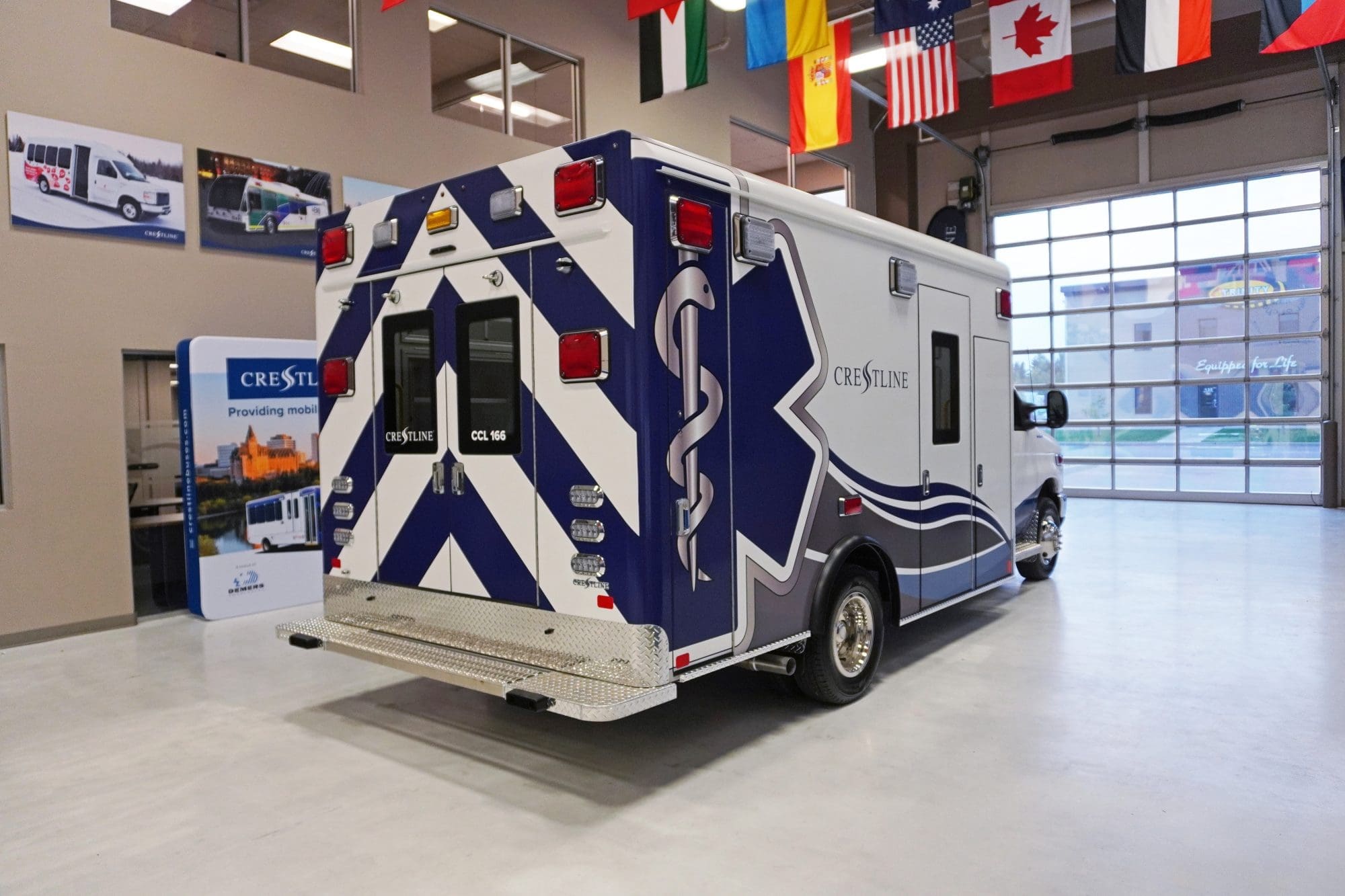 Rear and side view of a Crestline CCL 166 ambulance featuring a blue and white chevron design with the Star of Life graphic, photographed inside the Crestline manufacturing facility showroom