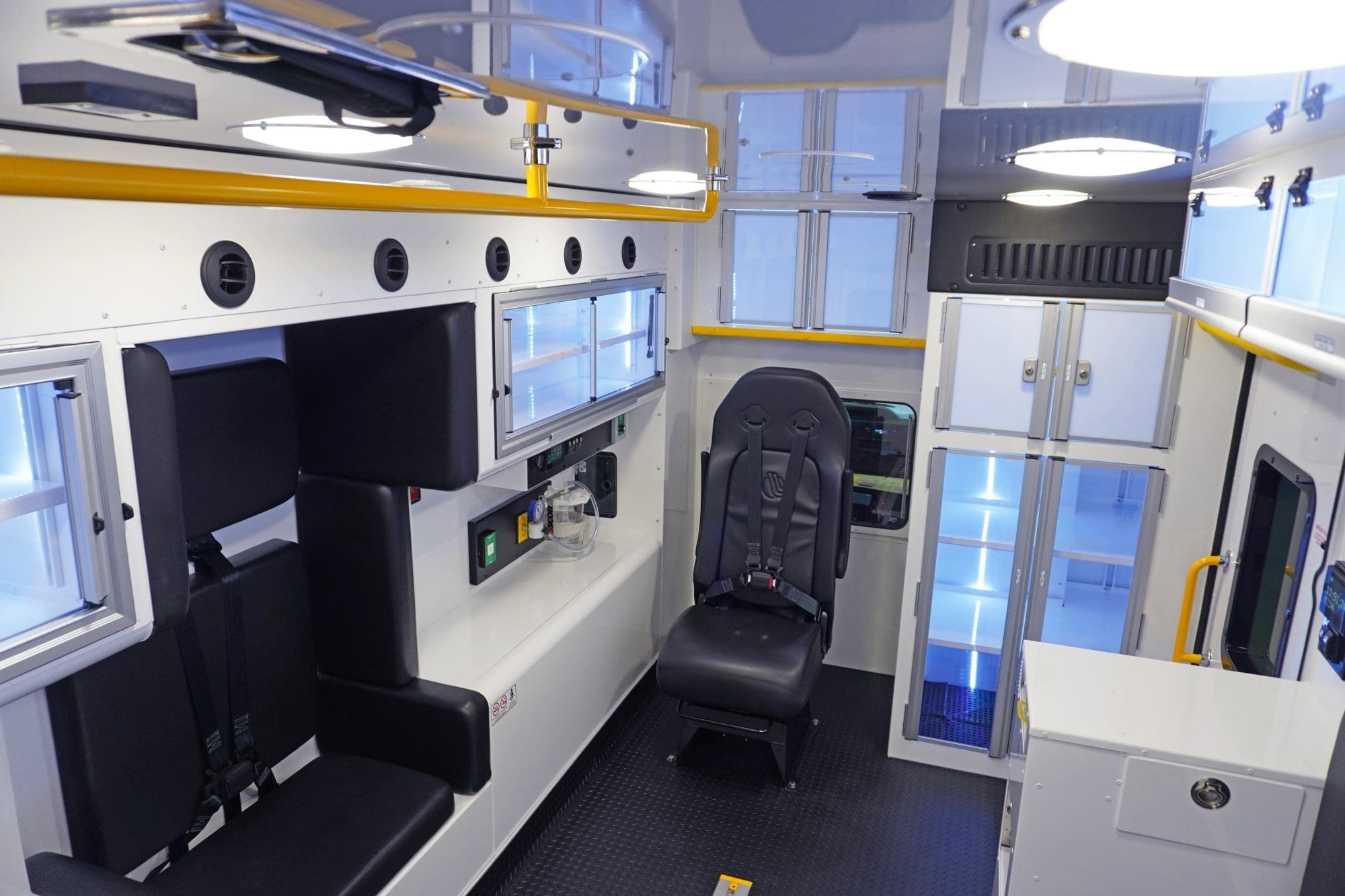 Wide interior view of a Crestline ambulance featuring black seating with seatbelts, upper and lower storage cabinets, medical equipment controls, and bright yellow grab handles