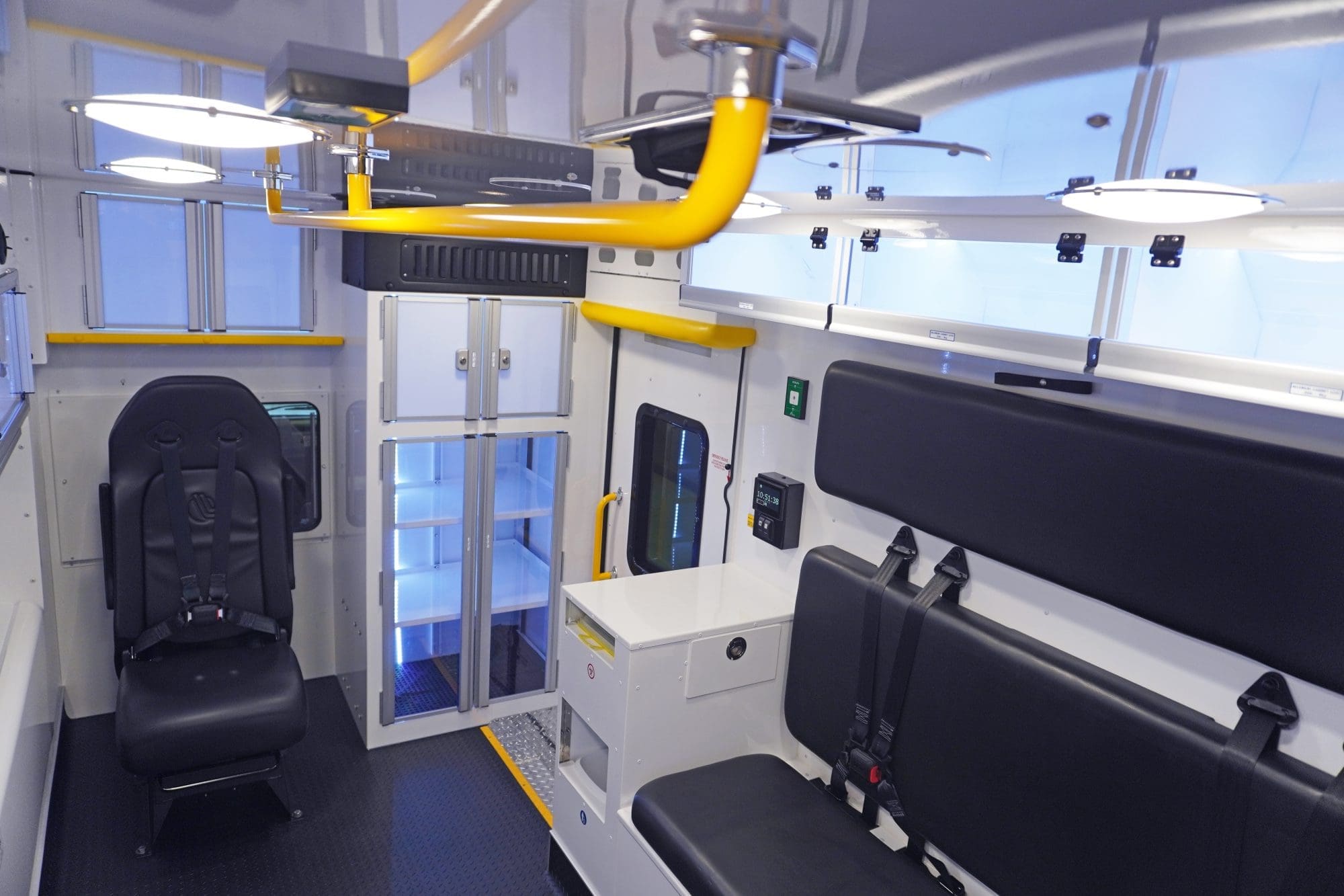 Wide view of a Crestline ambulance interior featuring black seating with safety harnesses, illuminated storage cabinets, yellow grab handles, and overhead lighting