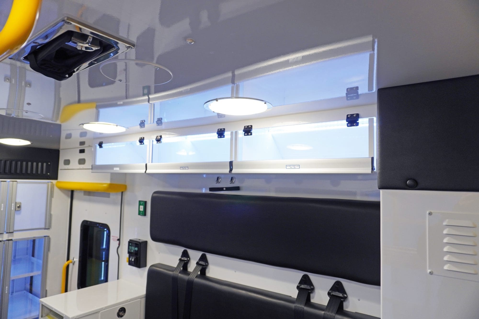 Overhead view inside a Crestline ambulance showing upper storage cabinets with clear doors, ceiling lighting, yellow grab handles, and a black bench seat below