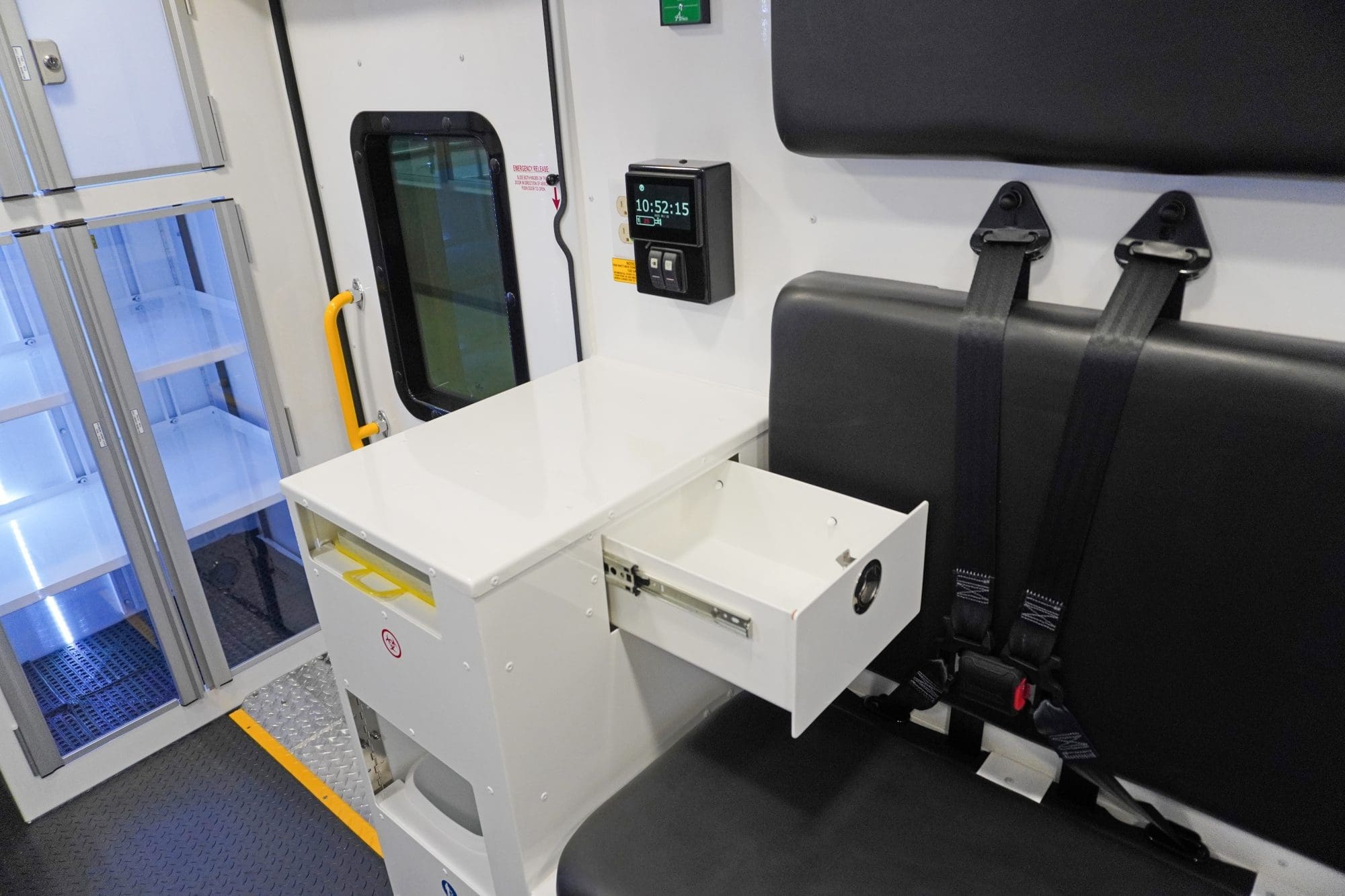 Crestline ambulance interior featuring a black bench seat with seatbelts, an open storage drawer above a biohazard waste unit, and illuminated supply cabinets near the side door