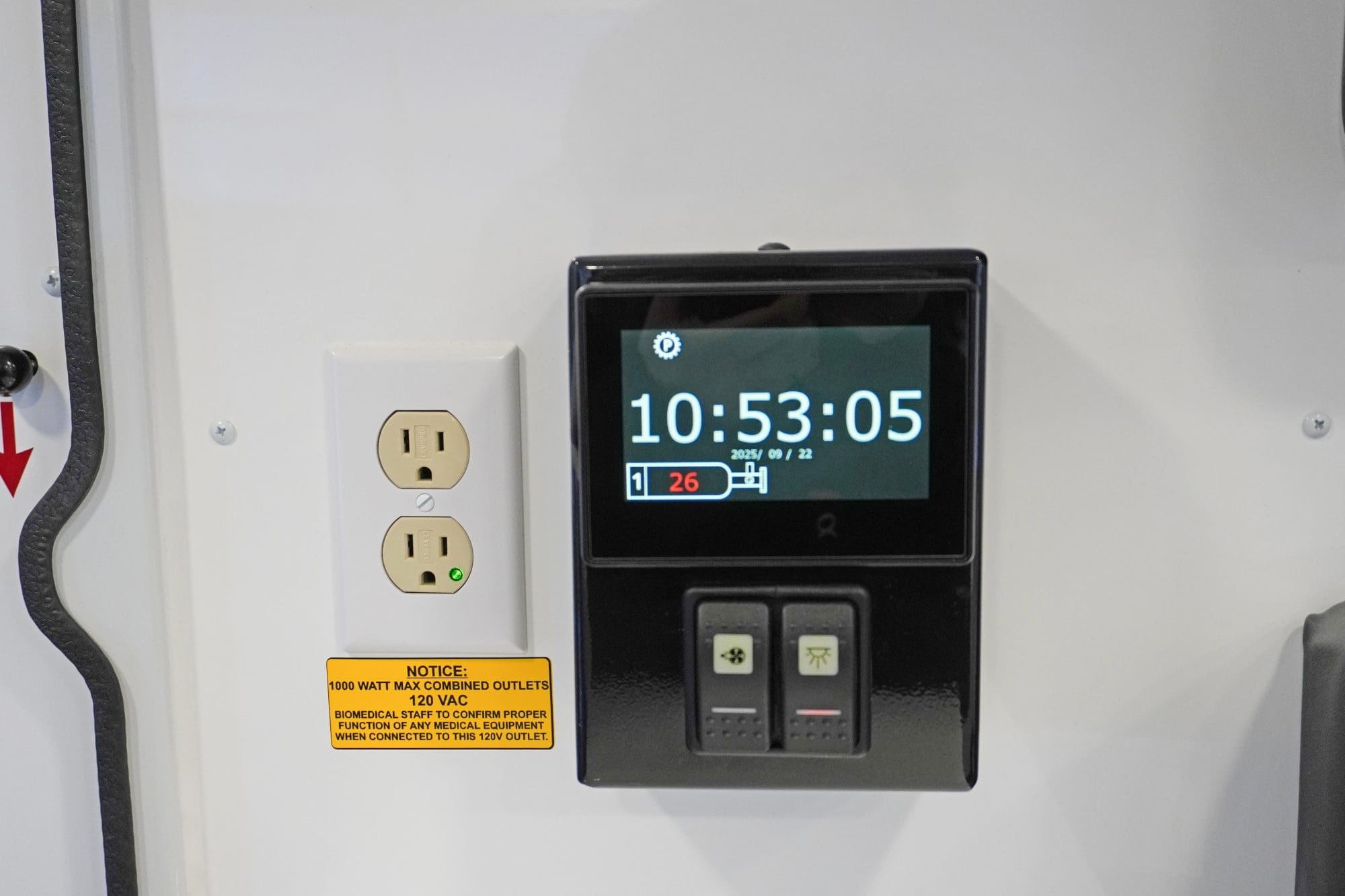 Close-up view of a Crestline ambulance interior wall panel featuring a digital control screen displaying the time and battery status beside a dual 120V power outlet with a caution label